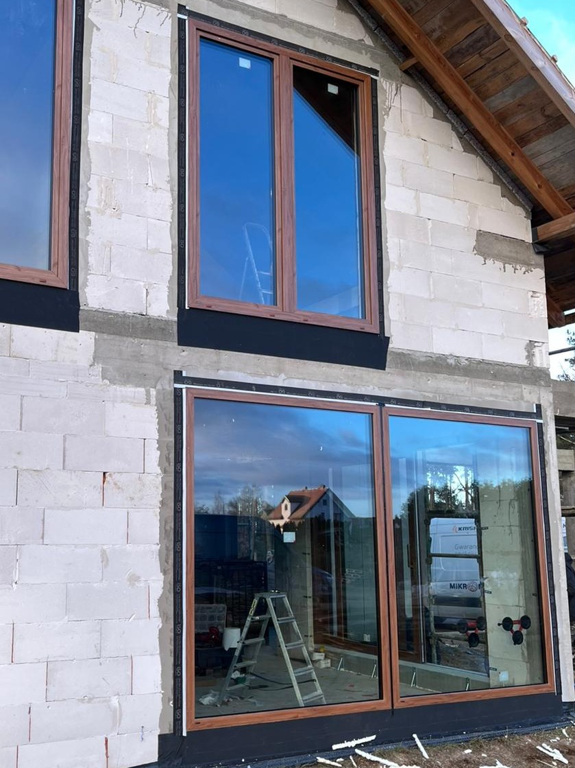 Montaż okien z brązowymi ramami w surowym budynku z białego betonu komórkowego, widoczna drabina i przyssawki do szyb.