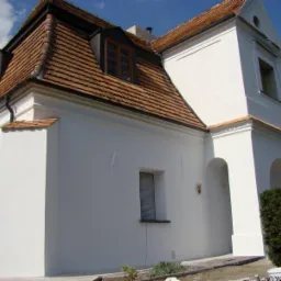 Odnowiony, biały dwór z czerwoną dachówką, widoczne okna dachowe i fragment ogrodu, wiadro z farbą przy ścianie.