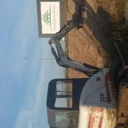 Mała koparka Bobcat 328 na placu budowy, z widocznym znakiem reklamowym Dunapack Eurobox w tle, pod błękitnym niebem.