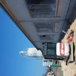 Mini koparka Bobcat obok wykopu pod fundamenty budynku z zabezpieczonymi oknami, widoczny silos w tle.