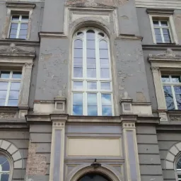 Fasada kamienicy z oknami w stylu klasycystycznym, widoczne uszkodzenia tynku i detale architektoniczne. W oknach odbija się błękitne niebo.