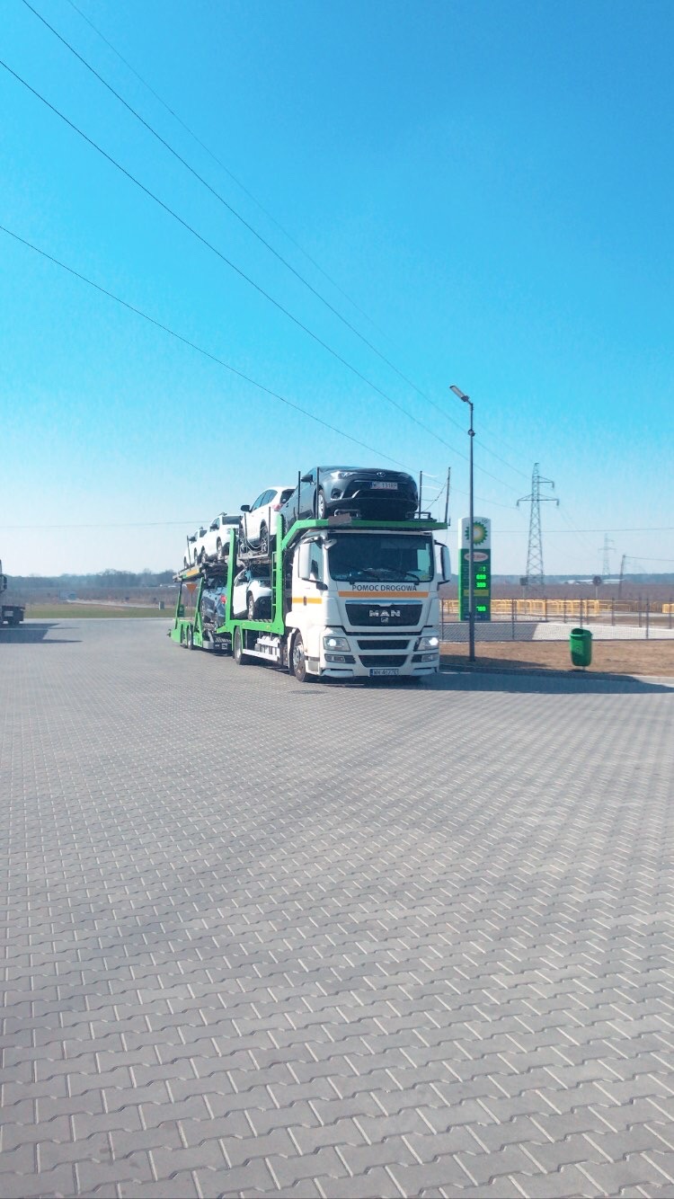 Laweta MAN z załadowanymi samochodami na parkingu stacji benzynowej w słoneczny dzień.
