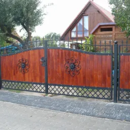 Drewniana brama wjazdowa i furtka z metalowymi elementami dekoracyjnymi w kształcie kwiatów, na tle drewnianego domu z ogrodem.