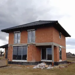 Dom w trakcie budowy z zamontowanymi oknami z białymi profilami i roletami zewnętrznymi, widoczne materiały budowlane i skrzynia na narzędzia.