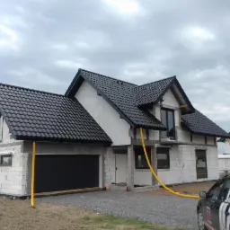 Surowy stan domu jednorodzinnego w trakcie budowy, z widoczną czarną bramą garażową, oknami i instalacją w żółtej rurze, na tle pochmurnego nieba.