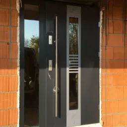 Nowoczesne, grafitowe drzwi wejściowe z długim, pionowym uchwytem i przeszkleniem, zamontowane w surowej ścianie z czerwonej cegły, widoczne ślady zaprawy.