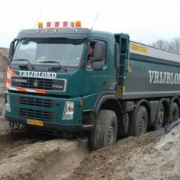 Ciężarówka wywrotka marki Volvo z ładunkiem porusza się po wykopie w piaszczystym terenie. Widoczny operator w kabinie. Na karoserii i kabinie widnieje napis 'VRIJBLOED'.