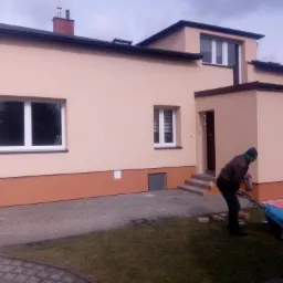 Dwukondygnacyjny dom murowany z ocieploną i pomalowaną elewacją w odcieniach beżu i brązu; robotnik w trakcie prac przygotowawczych, transportujący materiały budowlane wózkiem.