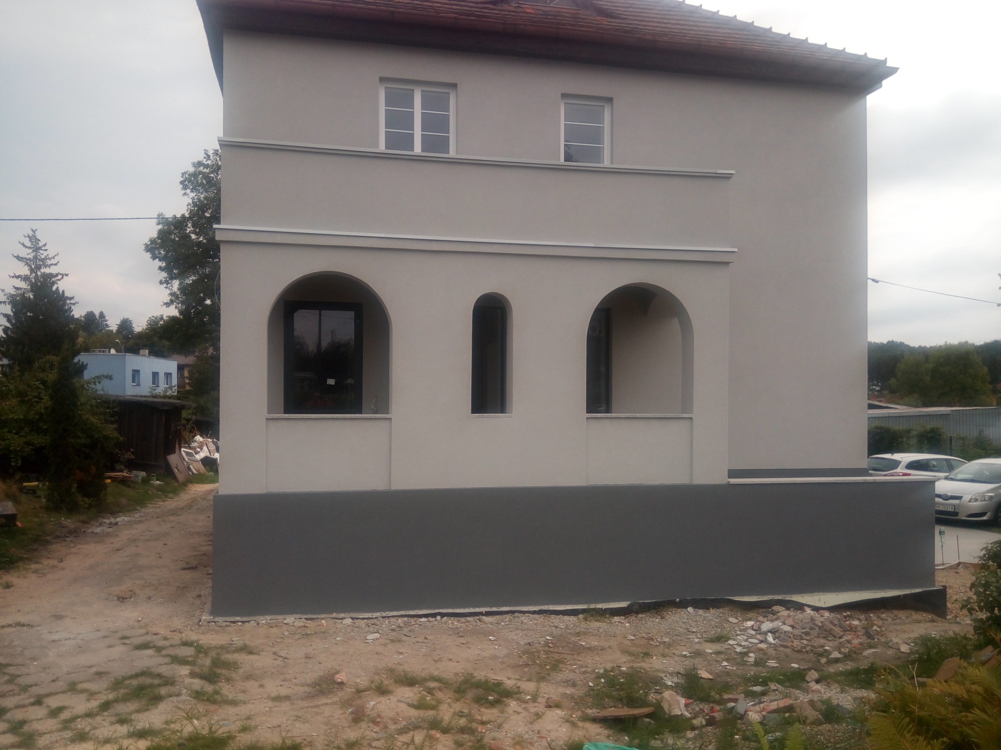 Dwukondygnacyjny dom z jasną elewacją i ciemnoszarą podmurówką, widoczne łukowate wnęki na parterze, teren wokół domu w trakcie prac wykończeniowych.