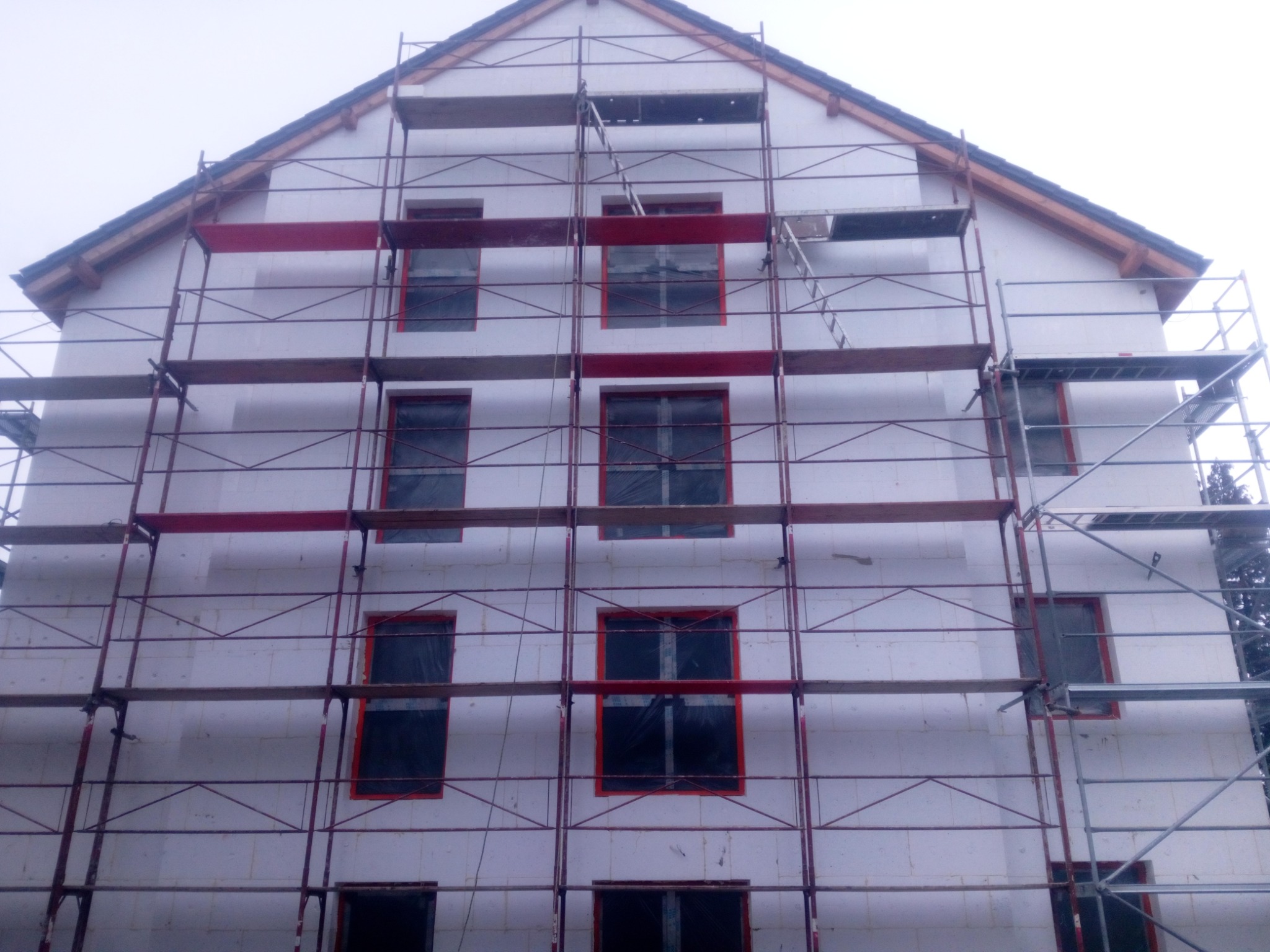 Dwupiętrowy dom w trakcie budowy z białych bloczków, z oknami zabezpieczonymi folią i czerwonymi obramowaniami, otoczony metalowym rusztowaniem.
