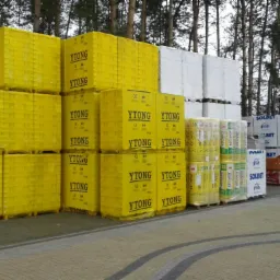 Składowane palety z bloczkami Ytong, wełną mineralną Isover i bloczkami Solbet na placu budowy, w tle drzewa. Widoczne pakiety materiałów budowlanych.