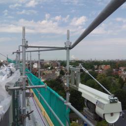 Grand World Company Sp.zo.o. - Widok z rusztowania na miasto, zabezpieczonego zieloną siatką i wyposażonego w podnośnik z napędem elektrycznym.