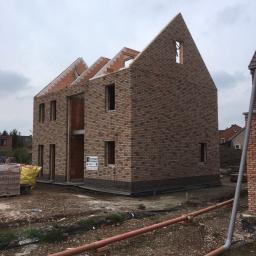 Grand World Company Sp.zo.o. - Dwukondygnacyjny dom w trakcie budowy z cegły klinkierowej, widoczne niezadaszone poddasze, okna bez ram, rury kanalizacyjne na ziemi.