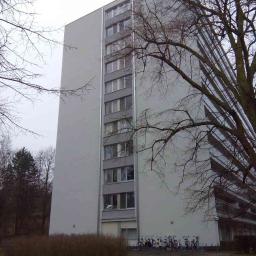 Grand World Company Sp.zo.o. - Wysoki, szary blok mieszkalny z balkonami, widoczny z perspektywy trawnika z zaparkowanymi rowerami i drzewami bez liści.