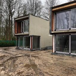 Grand World Company Sp.zo.o. - Dwa nowoczesne, dwukondygnacyjne domy w trakcie budowy, z dużymi przesuwnymi oknami i surowymi ścianami zewnętrznymi, otoczone błotnistym terenem budowy i drzewami bez liści.