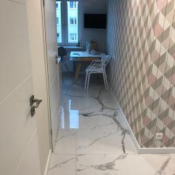 Grand World Company Sp.zo.o. - Otwarta biała płycinowa ościeżnica drzwiowa, widok na wnętrze z marmurową podłogą, geometryczną tapetą i częściowo widocznym stołem jadalnym.