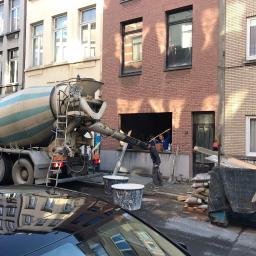 Grand World Company Sp.zo.o. - Betonomieszarka pompuje beton do otworu w ścianie budynku z czerwonej cegły, widoczni robotnicy wewnątrz, na pierwszym planie pojemniki i gruz.