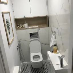 Nowoczesna toaleta z marmurowymi płytkami, geometryczną podłogą, wiszącą szafką i dekoracyjnymi elementami, takimi jak obraz z motywem roślinnym i pluszowe misie.