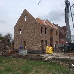 Grand World Company Sp.zo.o. - Wznoszenie dwukondygnacyjnego domu z cegły klinkierowej, widoczna konstrukcja dachu, palety z cegłami, zaprawa w workach i dźwig budowlany w tle.