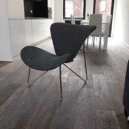 Grand World Company Sp.zo.o. - Szary, designerski fotel na metalowych nóżkach z kółkami na tle nowoczesnej kuchni z białymi szafkami i drewnianą podłogą.