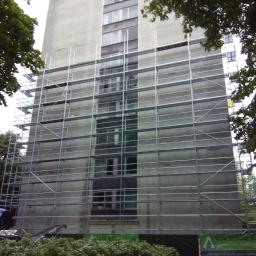 Grand World Company Sp.zo.o. - Wysoki budynek mieszkalny w trakcie renowacji, otoczony rusztowaniami, z widocznym fragmentem tablicy informacyjnej firmy budowlanej na pierwszym planie.