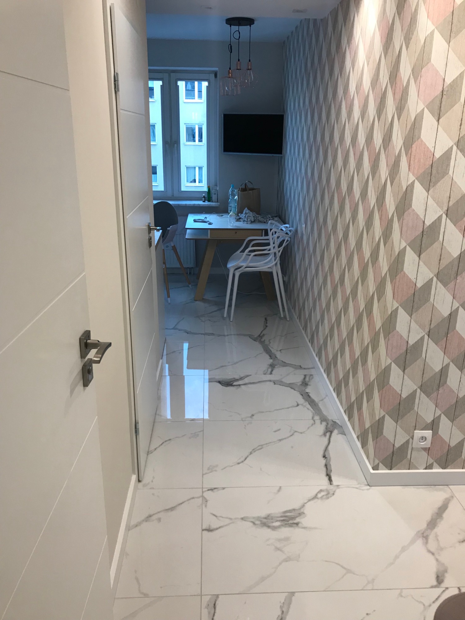 Otwarta biała płycinowa ościeżnica drzwiowa, widok na wnętrze z marmurową podłogą, geometryczną tapetą i częściowo widocznym stołem jadalnym.