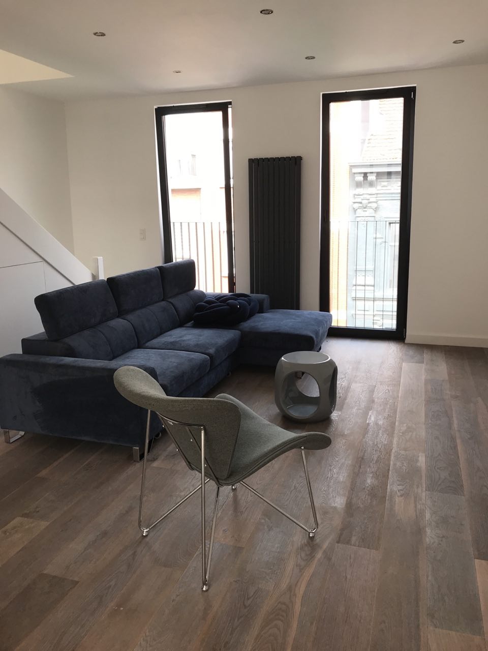 Jasne wnętrze pokoju dziennego z drewnianą podłogą, narożną sofą w odcieniach granatu, szarym krzesłem na metalowych nóżkach, grzejnikiem i dwoma oknami.