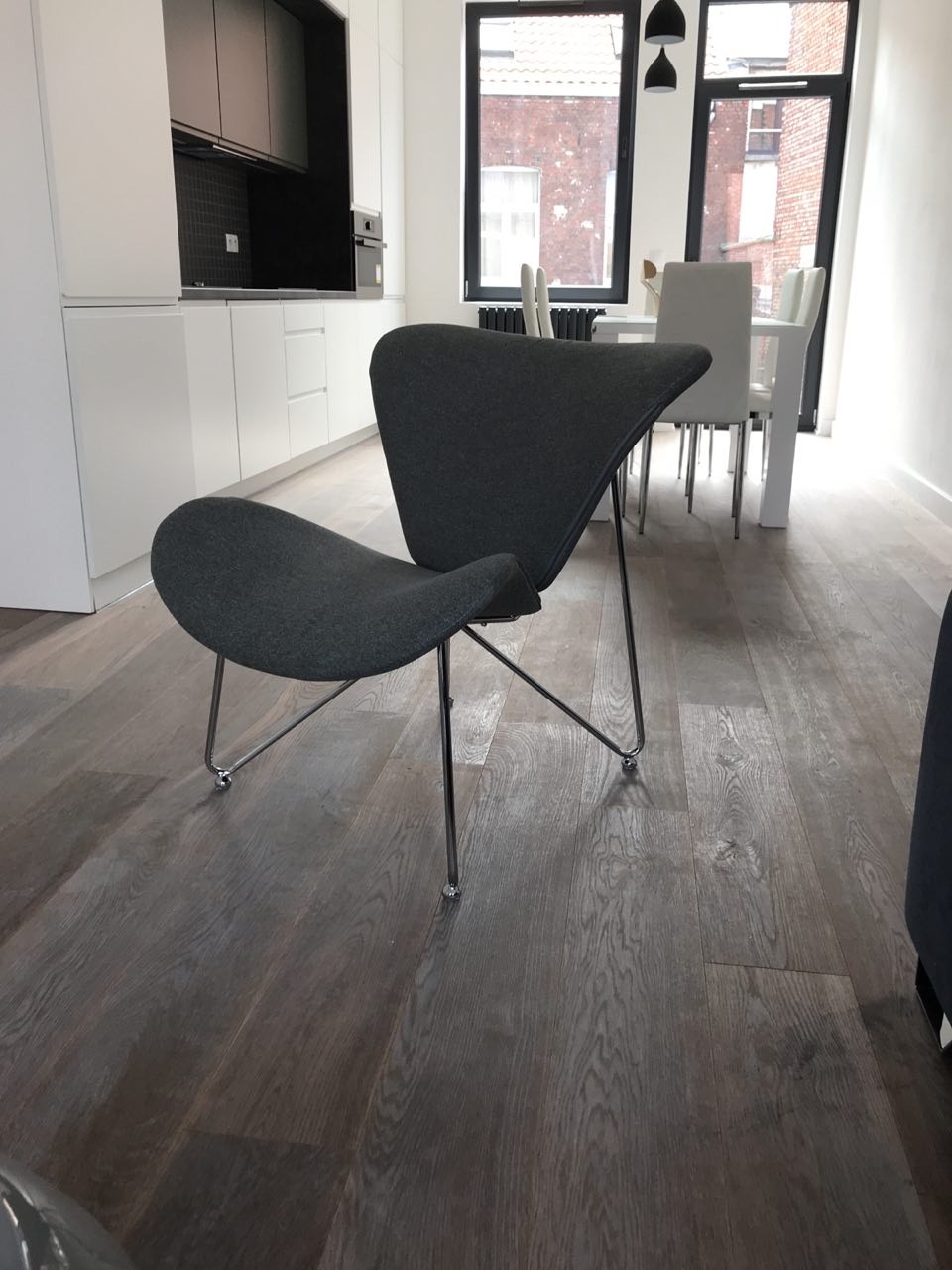 Szary, designerski fotel na metalowych nóżkach z kółkami na tle nowoczesnej kuchni z białymi szafkami i drewnianą podłogą.