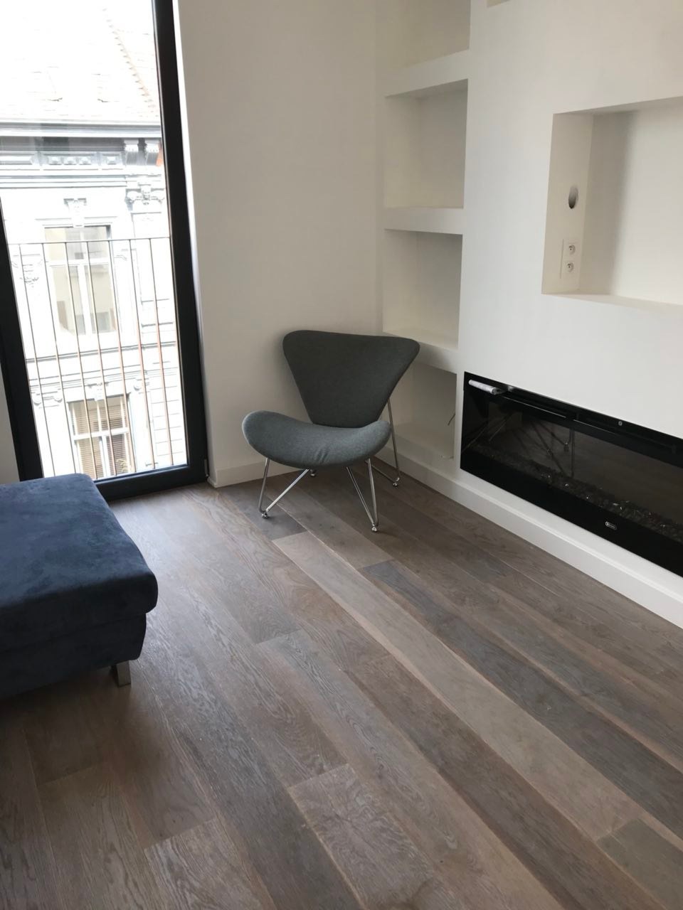 Jasne wnętrze z drewnianą podłogą ułożoną w jodełkę, szary fotel na metalowych nóżkach, zabudowany kominek elektryczny i widok na kamienicę za oknem.