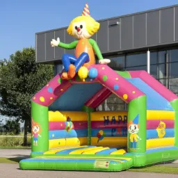 Kolorowy dmuchany zamek z postacią klauna w czapeczce siedzącego na szczycie, przed budynkiem z dużymi oknami.