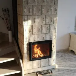 Piec kaflowy w odcieniach bieli, z palącym się wewnątrz ogniem, umieszczony w rogu pomieszczenia obok drewnianych schodów, z kawałkiem drewna leżącym na podłodze.