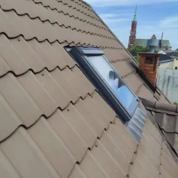 Beżowy dach z dachówki ceramicznej z zamontowanym oknem dachowym Velux; w tle widoczna wieża kościoła.