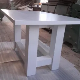 Biały, prostokątny stół z grubymi nogami, wykonany z drewna, sfotografowany w warsztacie na tle drewnianej podłogi i maszyn stolarskich.