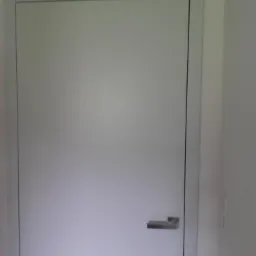 Zamknięte, białe drzwi wewnętrzne z prostokątną, metalową klamką, osadzone w białej futrynie, na tle jasnej ściany.