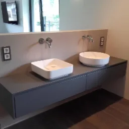 Nowoczesna łazienka z dwiema umywalkami nablatowymi: kwadratową i okrągłą, zamontowanymi na szarej, wiszącej szafce z dwiema szufladami i chromowanymi bateriami.