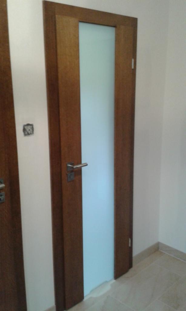 Drewniane drzwi wewnętrzne z matową szybą, widoczne z perspektywy narożnika pomieszczenia, z klamką i fragmentem drugiej pary drzwi po lewej stronie.