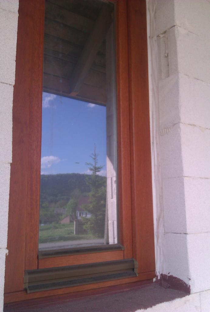 Wąskie okno z drewnianą ramą w surowym murze z widokiem na zalesione wzgórze i błękitne niebo.