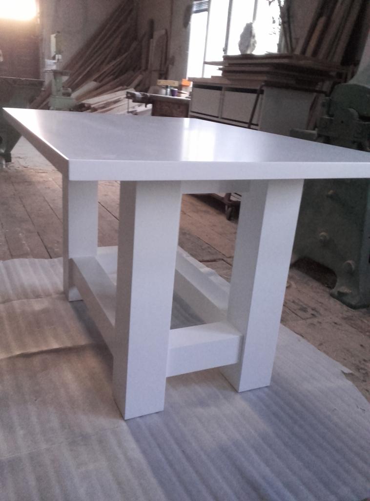 Biały, prostokątny stół z grubymi nogami, wykonany z drewna, sfotografowany w warsztacie na tle drewnianej podłogi i maszyn stolarskich.