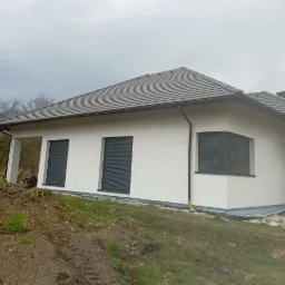 Nowoczesny dom jednorodzinny z białą elewacją i szarymi roletami zewnętrznymi, widok z boku, teren wokół domu częściowo uprzątnięty, widoczne ślady prac ziemnych.