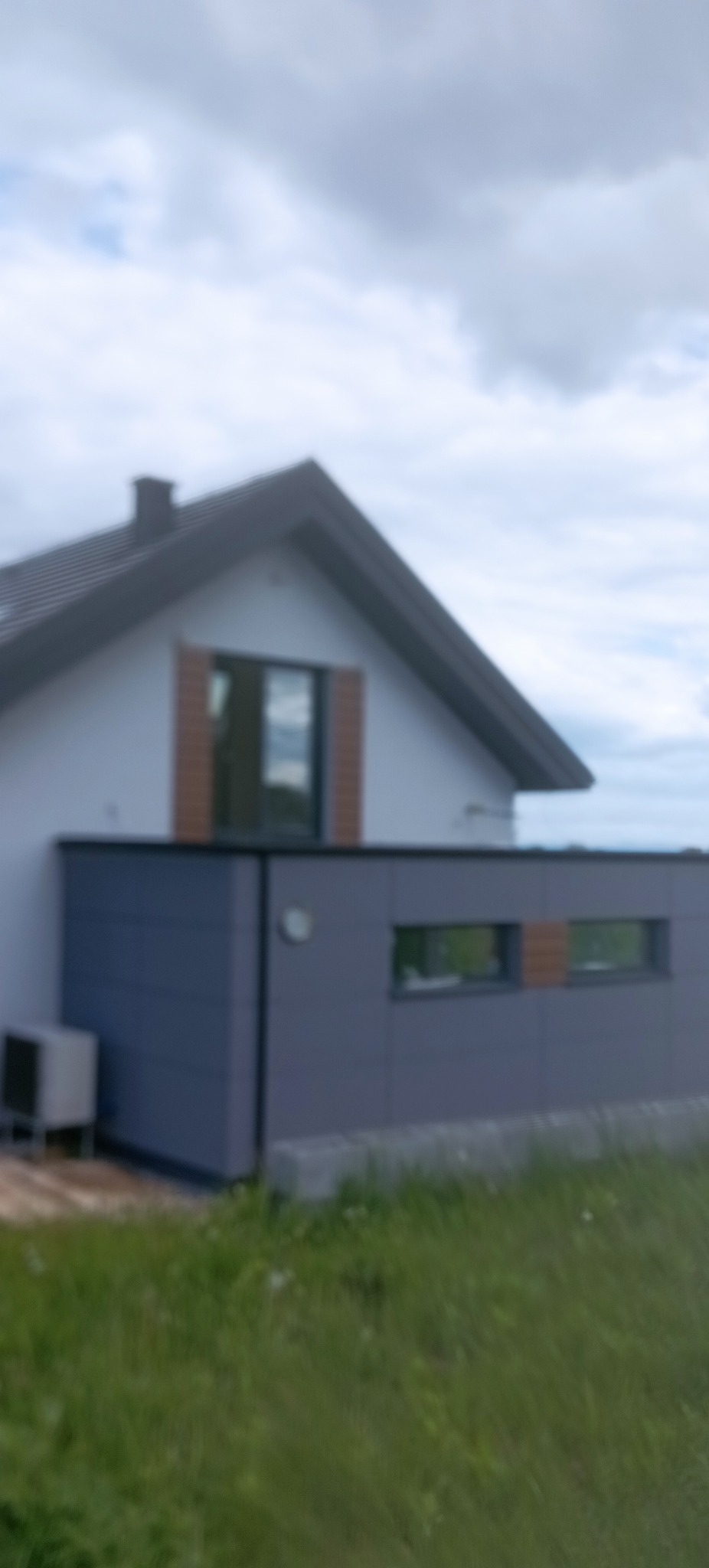 Dom jednorodzinny z szarą elewacją i brązowymi akcentami okiennymi, widok z zewnątrz, trawnik w pierwszym planie, pochmurne niebo w tle.