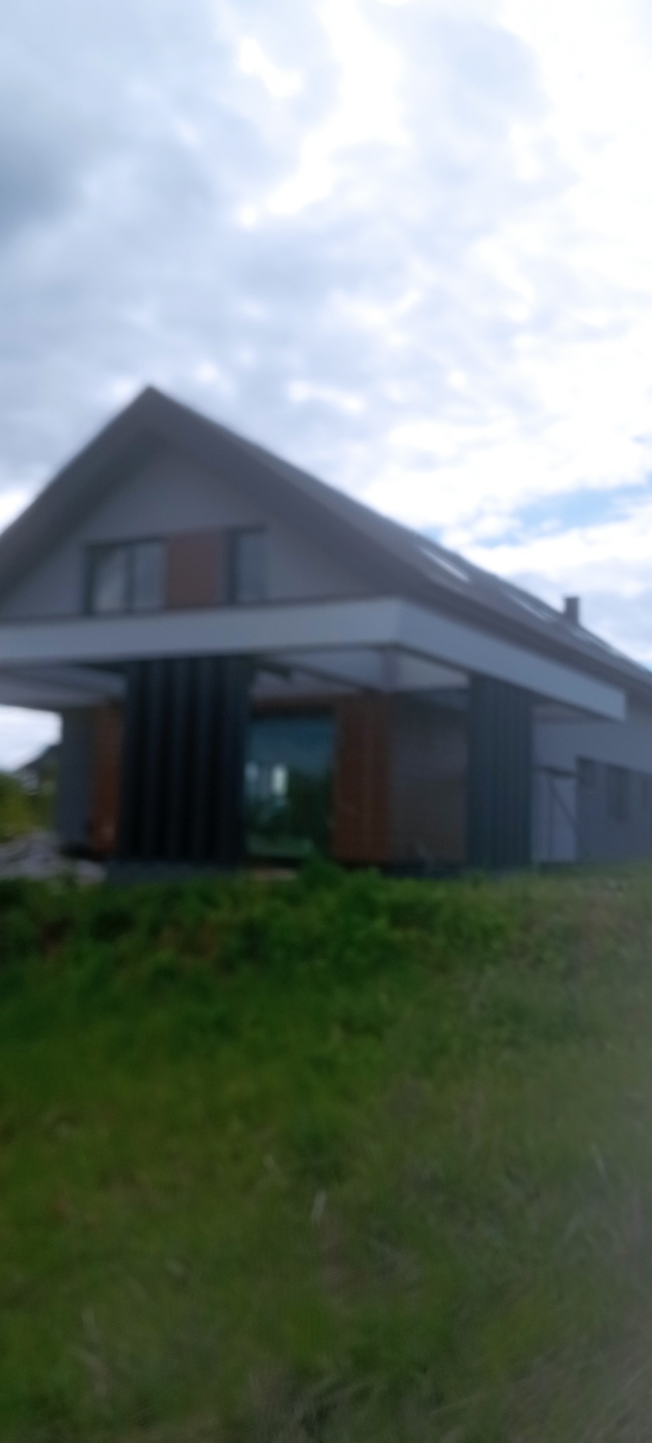 Nowoczesny dom jednorodzinny z szarą elewacją i elementami drewnianymi, widoczny z perspektywy trawnika. Pochmurne niebo w tle. Rozmycie obrazu.