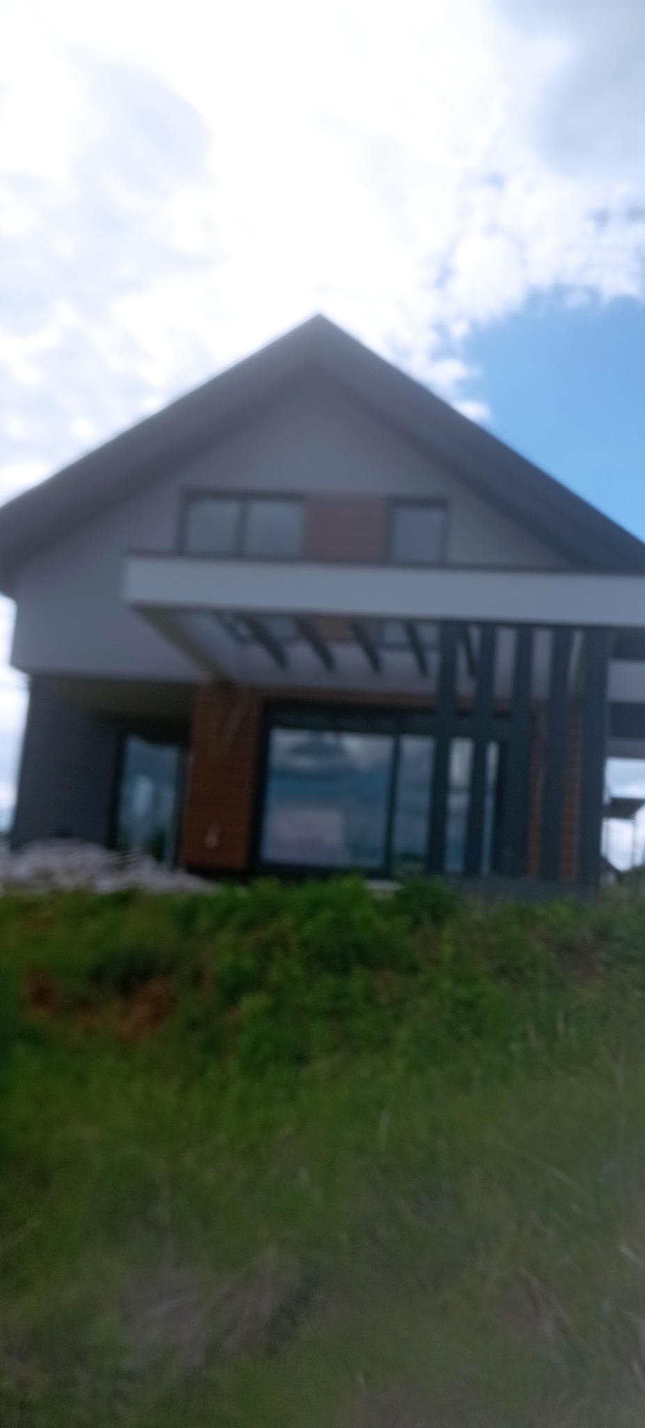 Nowoczesny dom jednorodzinny z szarą elewacją i drewnianymi akcentami, widoczny z perspektywy trawnika. Duże okna i minimalistyczny design.