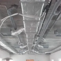 Widok na sufit z zainstalowaną instalacją wentylacyjną: prostokątne kanały owinięte folią aluminiową, okrągłe rury PCV, elastyczne przewody wentylacyjne typu spiro, skrzynki wentylacyjne i kable...