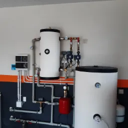 Zestaw dwóch białych zbiorników akumulacyjnych ciepła z podłączeniami hydraulicznymi, manometrami i zaworami, zamontowany na ścianie z pomalowanym poziomym pasem w kolorach szarym i pomarańczowym.