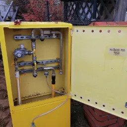 Otwarta, żółta skrzynka przyłącza gazowego z widocznymi rurami, zaworami i reduktorem, umieszczona na zewnątrz budynku, z butelką wody u dołu.