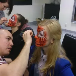 Kobieta maluje twarz dziewczynie w motywie Spidermana pędzelkiem, druga osoba z podobnym makijażem w tle, w pomieszczeniu z czarnymi krzesłami i lustrem.