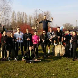 Grupa kilkunastu osób z kijkami do Nordic Walking pozuje do zdjęcia w parku, jeden z mężczyzn stoi na pniu drzewa z podniesionymi kijkami.