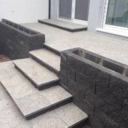 Schody i taras wyłożone jasną kostką brukową, obrzeża z ciemnej kostki. Widoczne donice z kostki na rośliny, nowoczesny design.