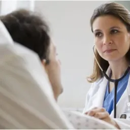 Lekarka w białym fartuchu, ze stetoskopem na szyi, bada pacjenta leżącego w łóżku szpitalnym.