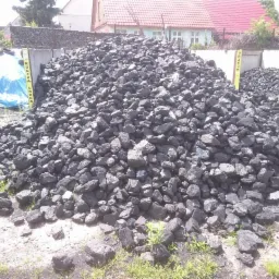 Góra czarnego węgla kamiennego na placu składowym, widoczne oznaczenia 'Kostka Gruba' i 'Orzech', w tle budynek z czerwoną dachówką i niebieska plandeka.